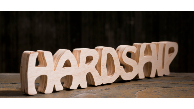 The word HARDSHIP is carved in bold, three-dimensional wooden letters, placed on a rustic wooden surface with a dark, blurred background.