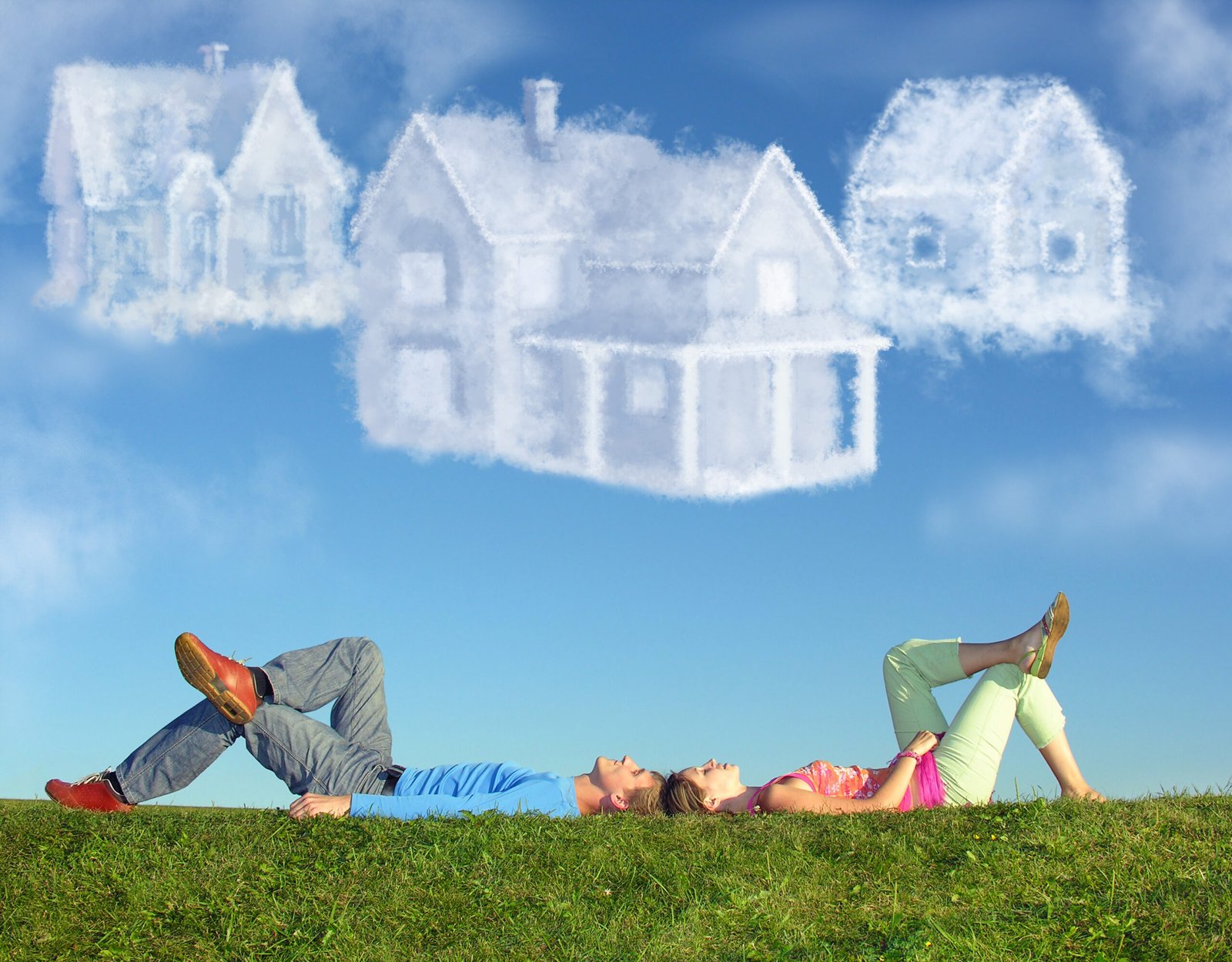 A man and woman lie on grass, looking up at the sky where three large, white cloud shapes resemble houses, symbolizing dreaming or planning for a future home.