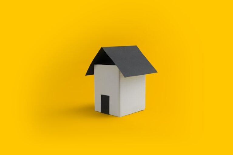 A small, minimalist model of a house with a black roof and door sits centered on a bright yellow background, evoking the modern appeal of a multifamily real estate sale in California.